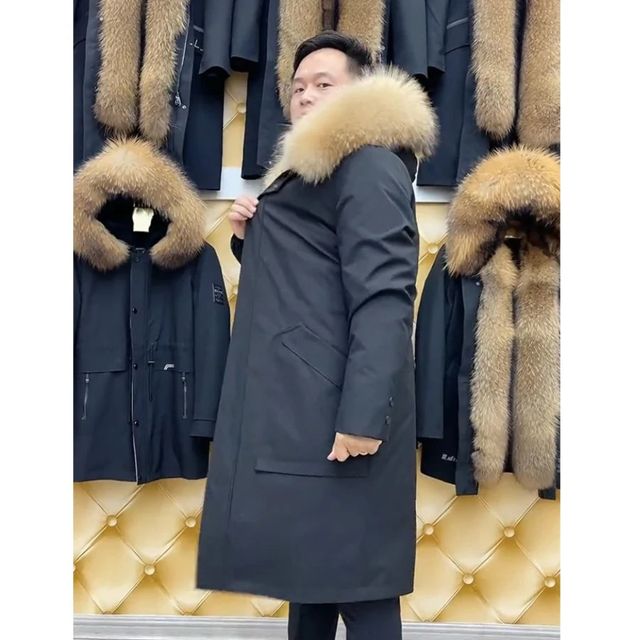 Side view of men's mid-calf coat in River Bed color with voluminous raccoon hood and detachable muskrat fur lining, showing straight silhouette and front patch pocket. From Borcelleclothes.