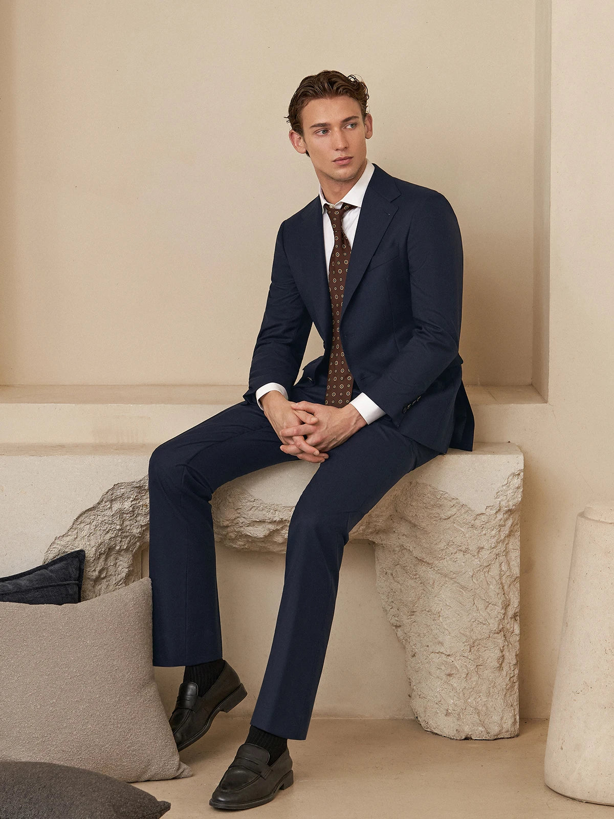 Model sitting in the Borcelleclothes Men's Ebony Clay Business Suit against a modern decorative stone backdrop, showcasing the suit's elegant tailoring and professional appeal.
