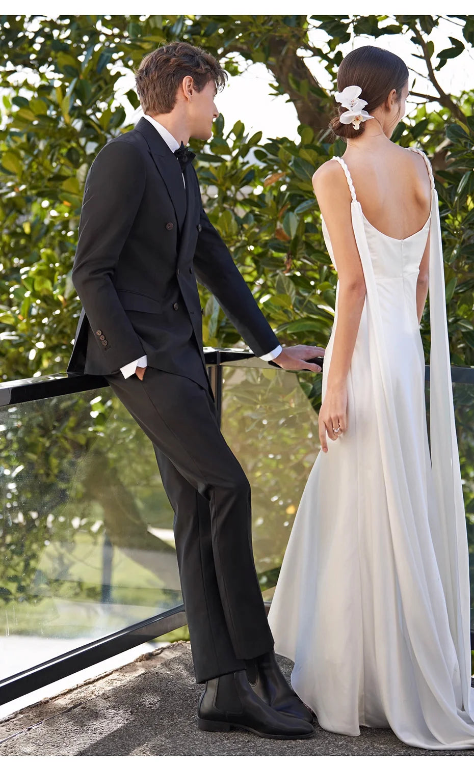 Model in Ebony Clay wool suit talking to a bride on a balcony — a moment that doesn't ask, but invites. From Borcelleclothes.