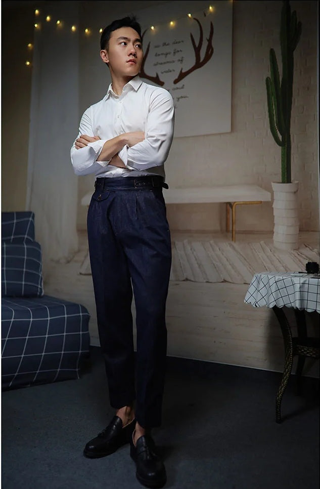 Model with arms crossed, wearing tailored denim trousers with front tucks and dual side buckles. From Borcelleclothes.