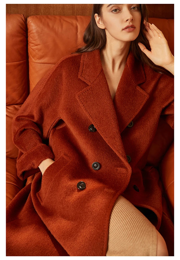 Model wearing the Women’s Mid-Calf Alpaca Coat in Cognac, seated on a sofa, showcasing the coat’s rich color and elegant drape. From Borcelleclothes.