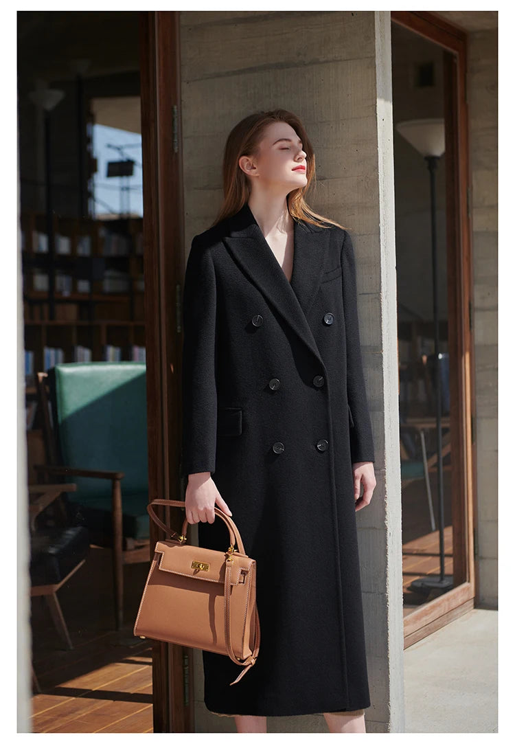 Model in an Onyx cashmere wool coat leaning softly against a neutral wall. From Borcelleclothes.