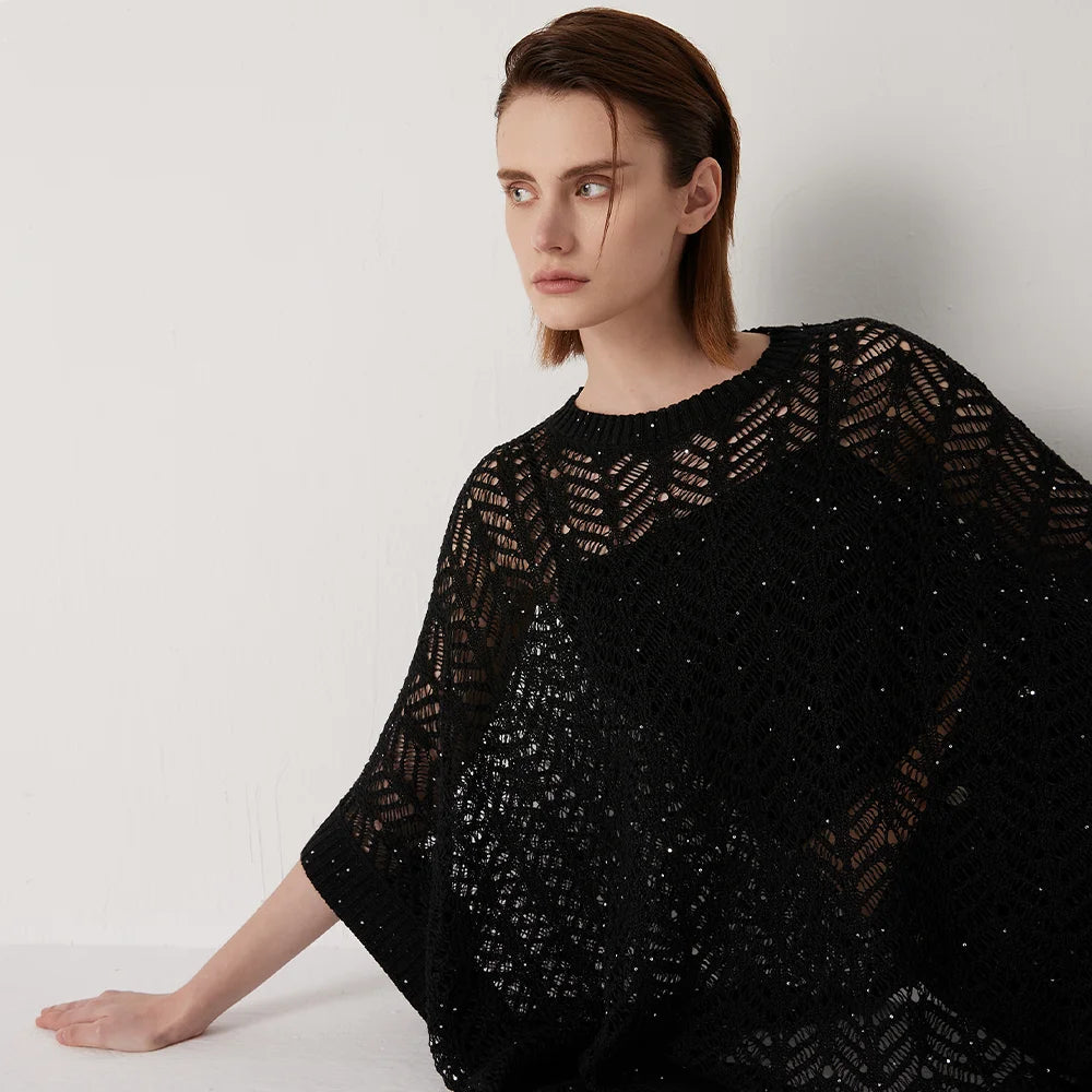 Model in Onyx lace poncho reclining slightly against a table, abstract openwork knit catching soft directional light. From Borcelleclothes.
