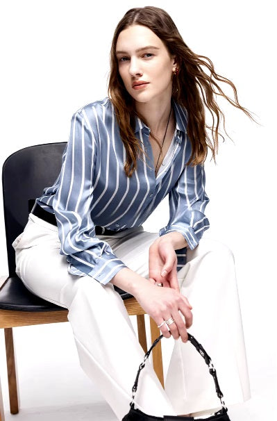 Model wearing Women's Luxury 6A Mulberry Silk Shirt in Striped Light Blue Grey & White, sitting on a chair, highlighting the relaxed fit and luxurious silk texture. From Borcelleclothes.