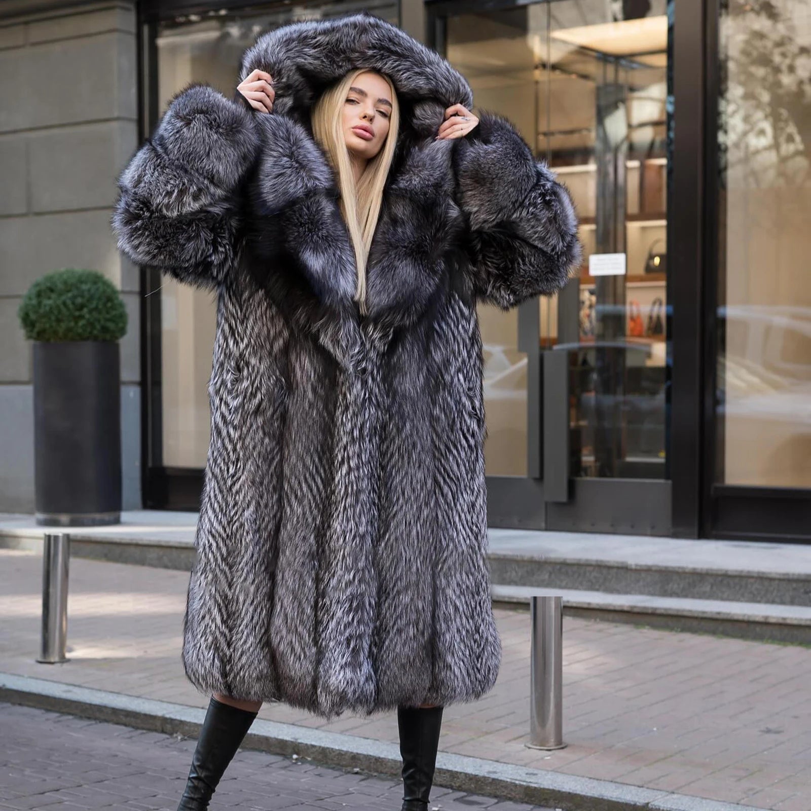 A model wearing a Women's Silver Fox Fur Coat, touching the lifted expanded hood, highlighting its dramatic silhouette and luxurious texture. From Borcelleclothes.
