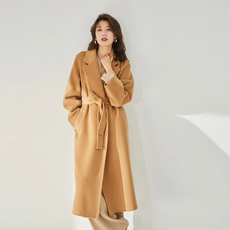 A model wearing the Women's Wrap Coat Redefined in Brown Sugar, gently touching the collar, emphasizing its refined notch lapels and tailored elegance. From Borcelleclothes.