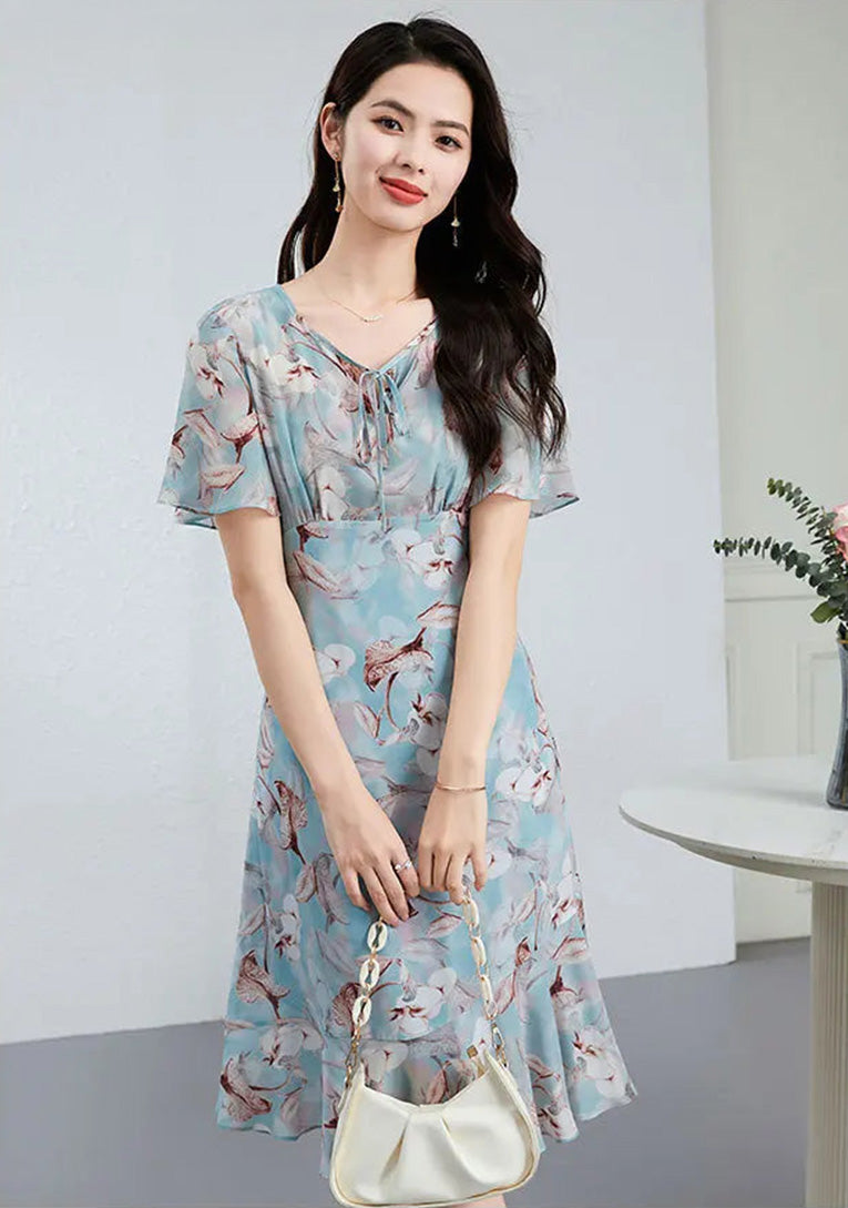 A woman in a flowing sky-blue silk dress, holding her handbag with both hands in a poised, elegant stance — From Borcelleclothes.