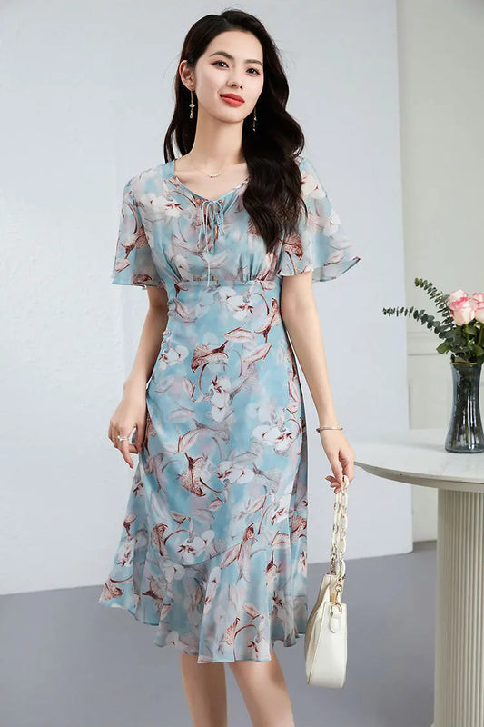 A woman in an asymmetrical silk empire waist dress in Celestial Bauhinia blue, gracefully holding a white structured bag — From Borcelleclothes.