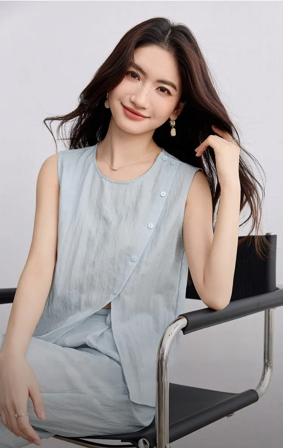 A woman sitting gracefully, touching her hair while wearing the light blue asymmetrical Lyocell top and wide-leg trousers — From Borcelleclothes.
