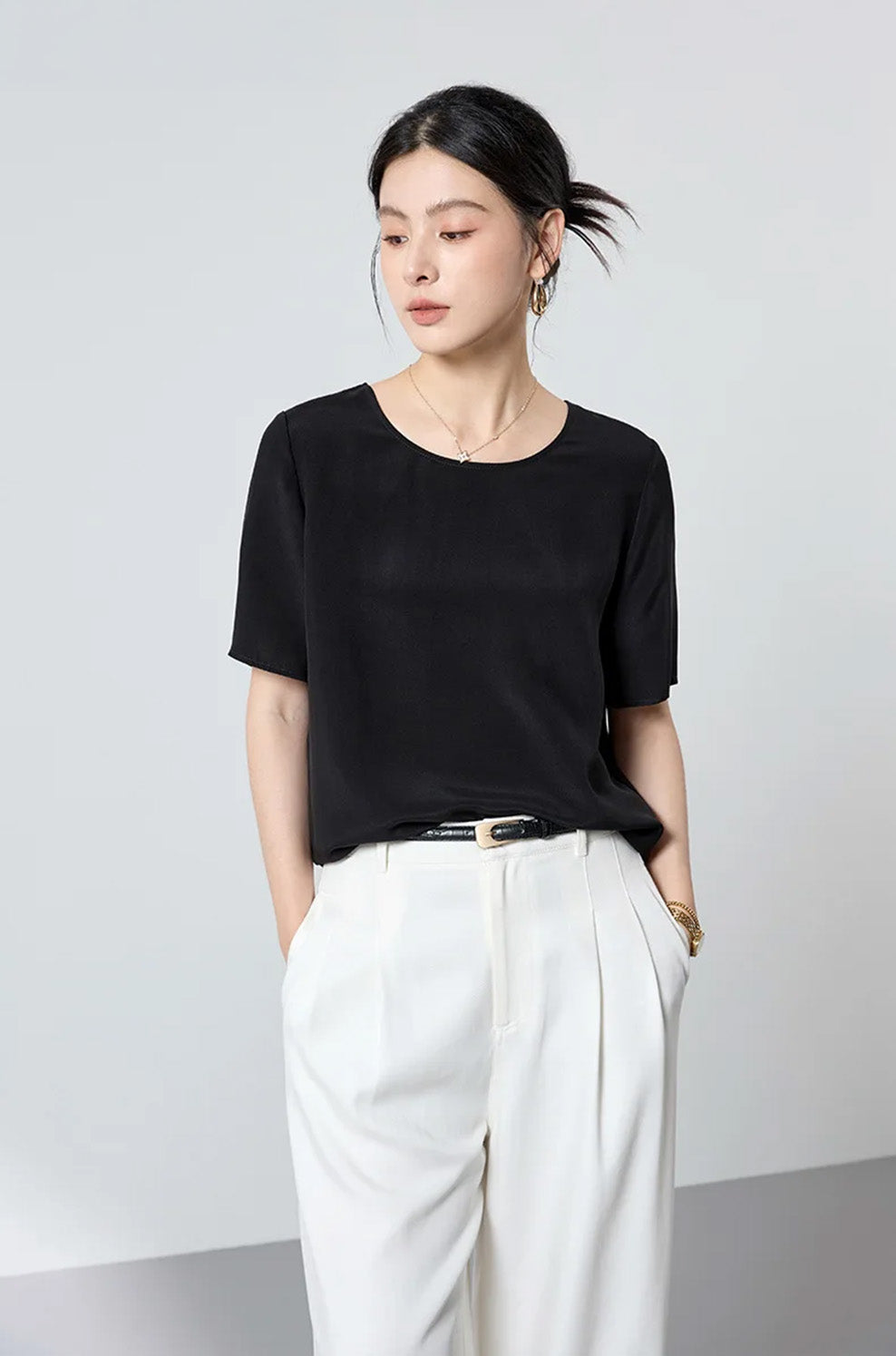 Model with both hands in the pockets of white tailored trousers, showing the clean drape and hip-length fit of the black silk-viscose top — From Borcelleclothes.
