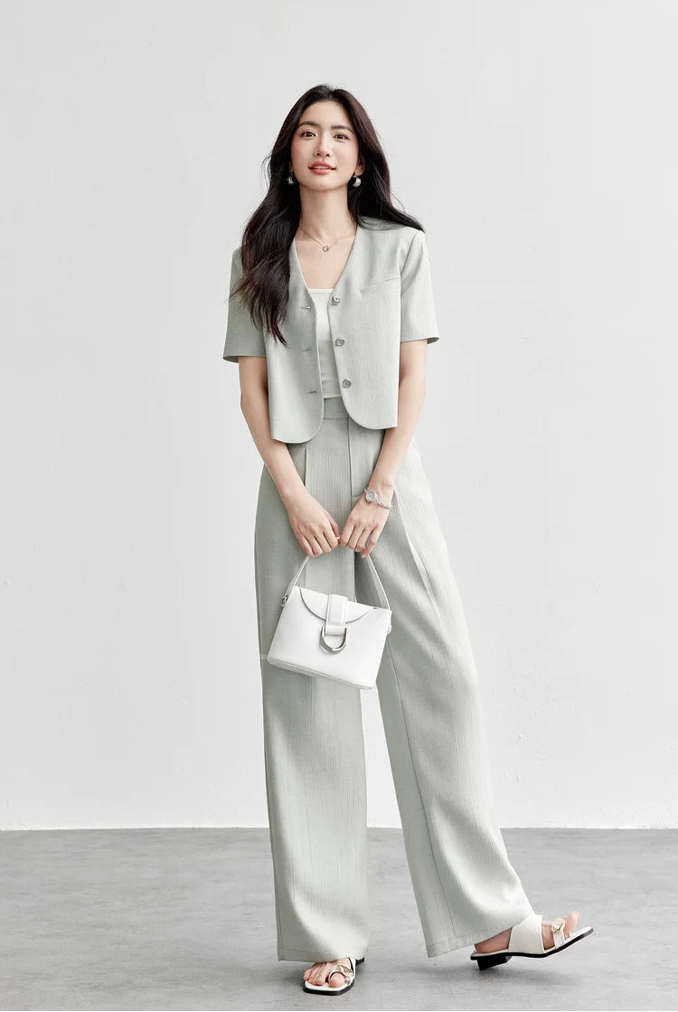 A woman in a sage-grey viscose set holding a minimalist white bag, showcasing the clean geometry of the cropped jacket and wide-leg trousers. From Borcelleclothes.