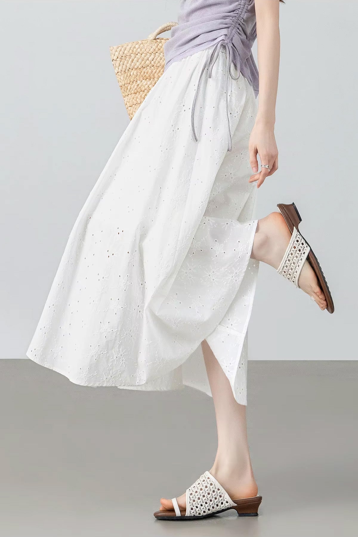 Dynamic shot of a model with a bent leg adjusting her sandal, revealing the flexible movement and midi length of the white eyelet skirt — From Borcelleclothes.