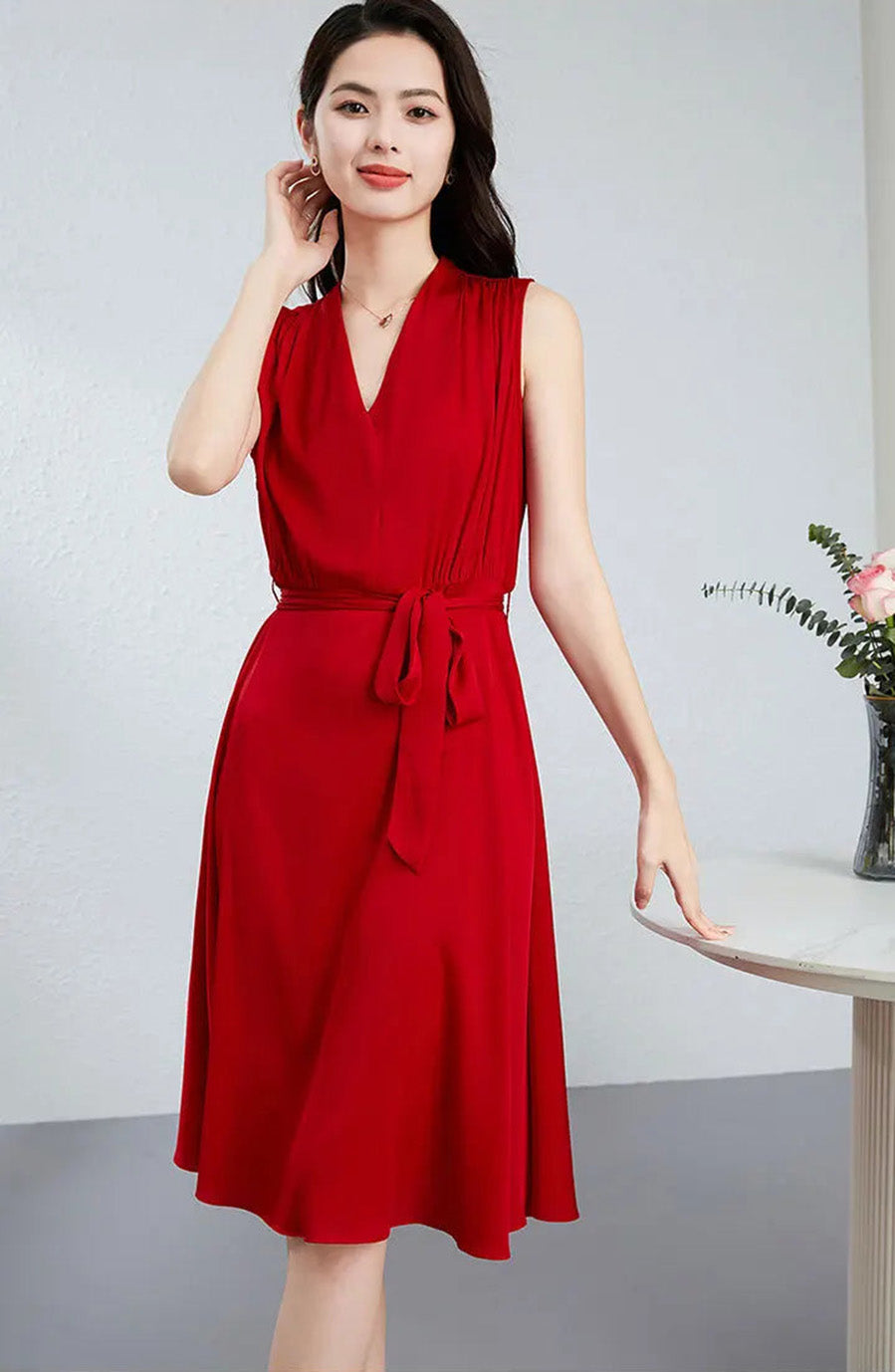 A woman adjusts her hair while wearing a flared sleeveless silk dress, revealing the clean-front bodice and architectural draping — From Borcelleclothes.