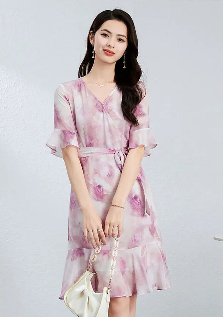 A woman in a flared silk wrap dress in Highland Heather, holding a white bag with both hands with calm confidence — From Borcelleclothes.