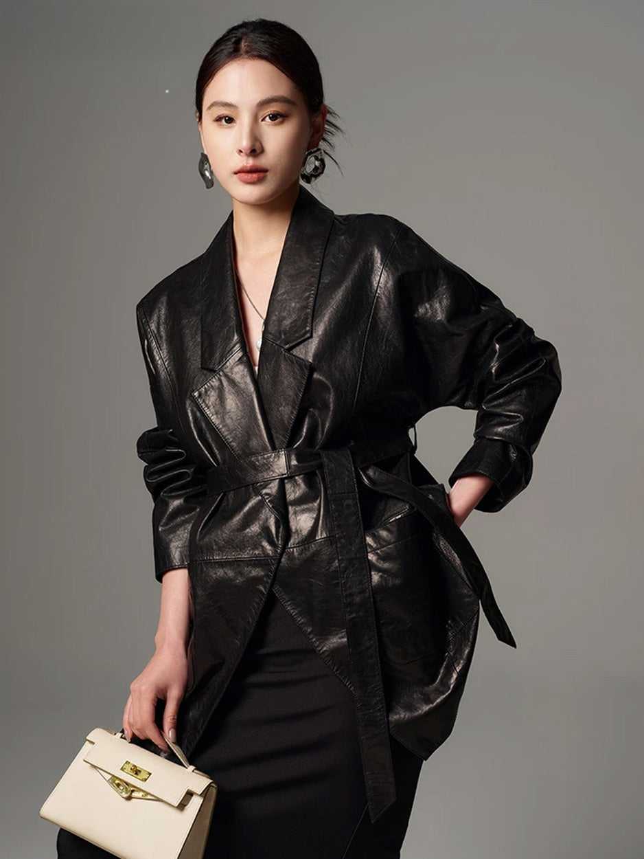 Model posing with a side-bent posture in the belted Vola Con Me glazed goatskin wrap jacket, holding a structured cream bag — From Borcelleclothes.