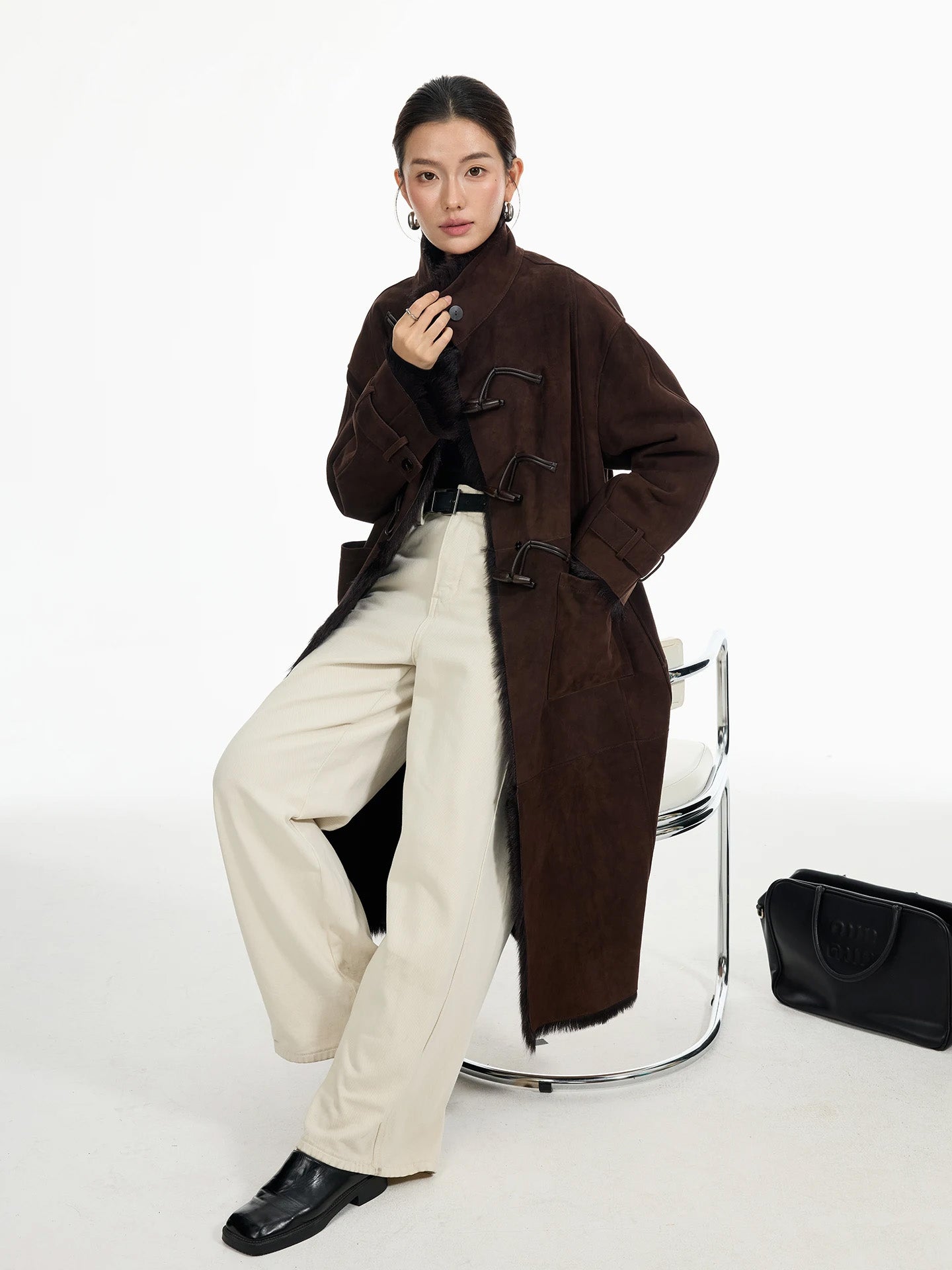 Model seated in goatskin coat, Wenge, touching wrap stand collar; leather air loops arranged diagonally across the front — From Borcelleclothes.
