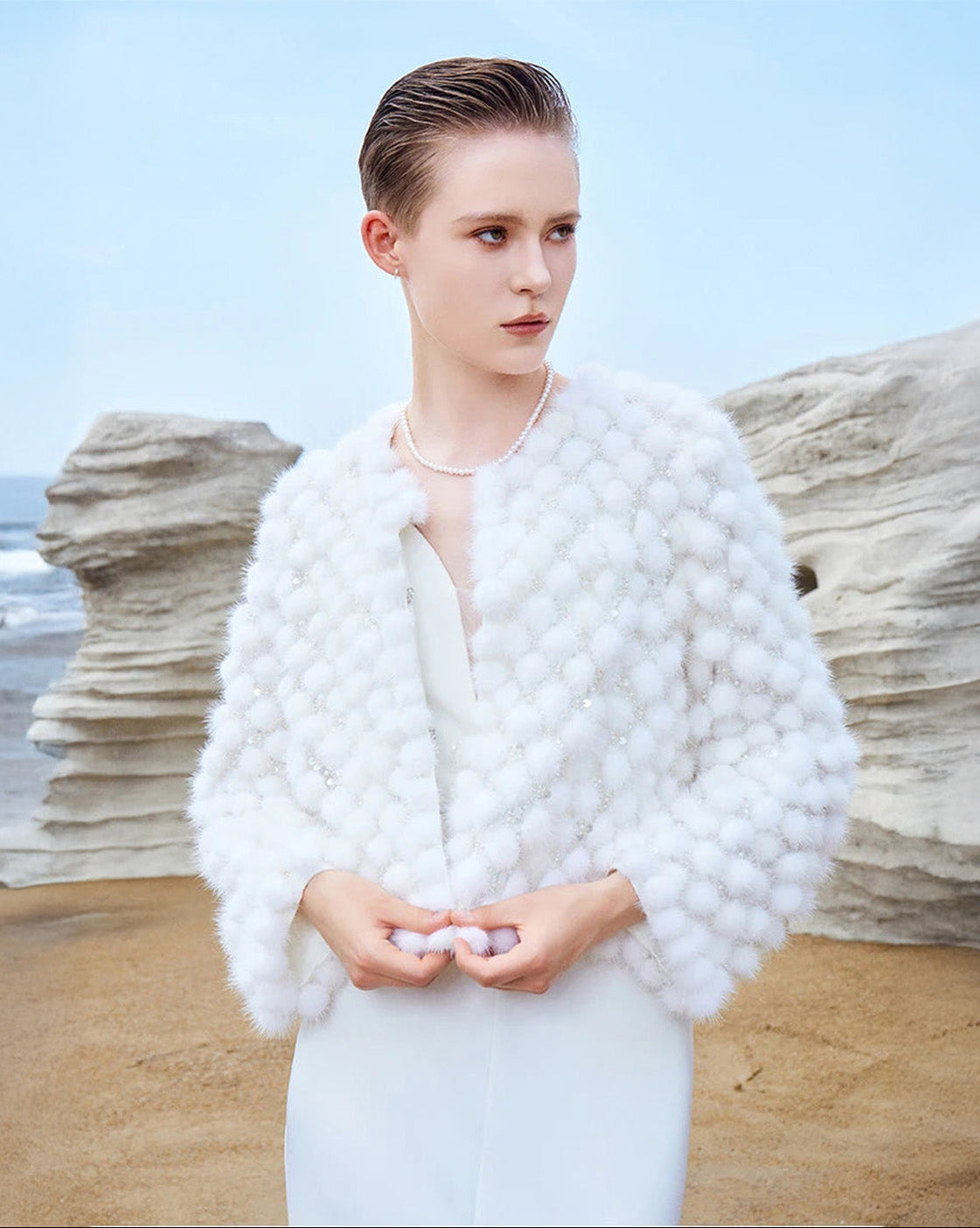 Model holding the bottom pom-poms of the Women’s Mink Mosaic Coat in White Whimsy, emphasizing handcrafted detail — From Borcelleclothes.