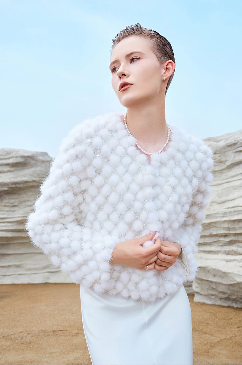 Model holding the front edges of the White Whimsy mink mosaic coat while looking to the side, showing fur detail and soft silhouette — From Borcelleclothes.