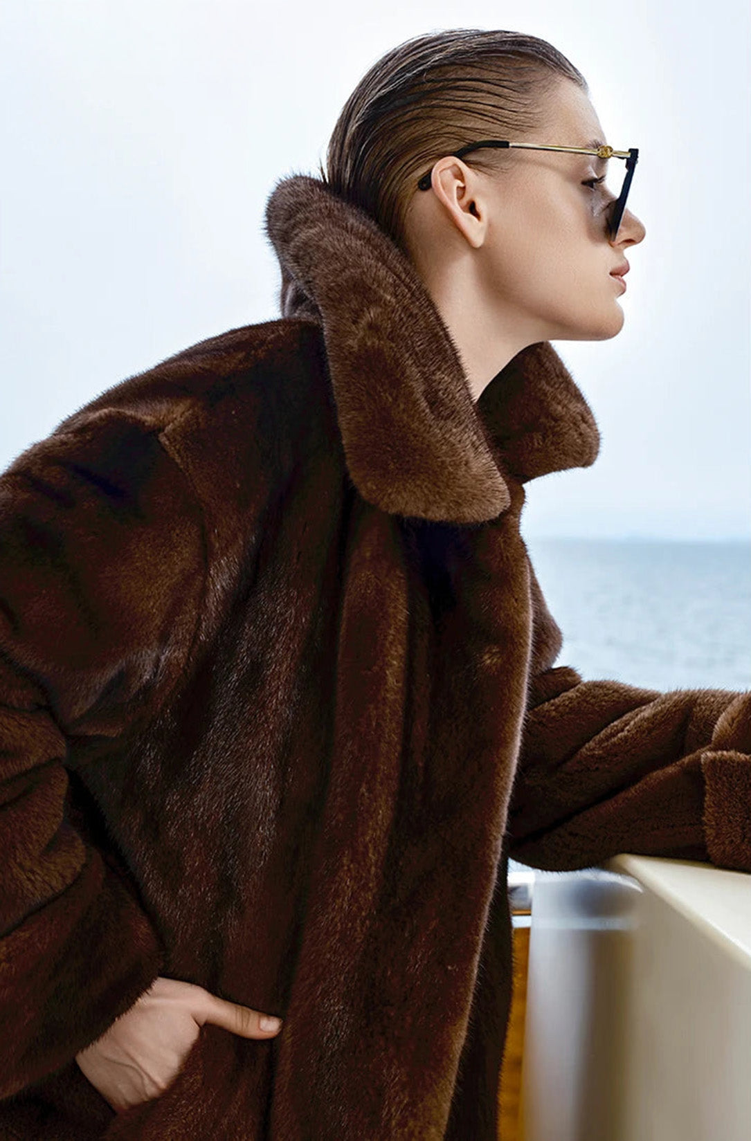 Model in Mirage of Marron mink coat, standing near yacht railing, gazing toward the sea in soft daylight. — From Borcelleclothes.