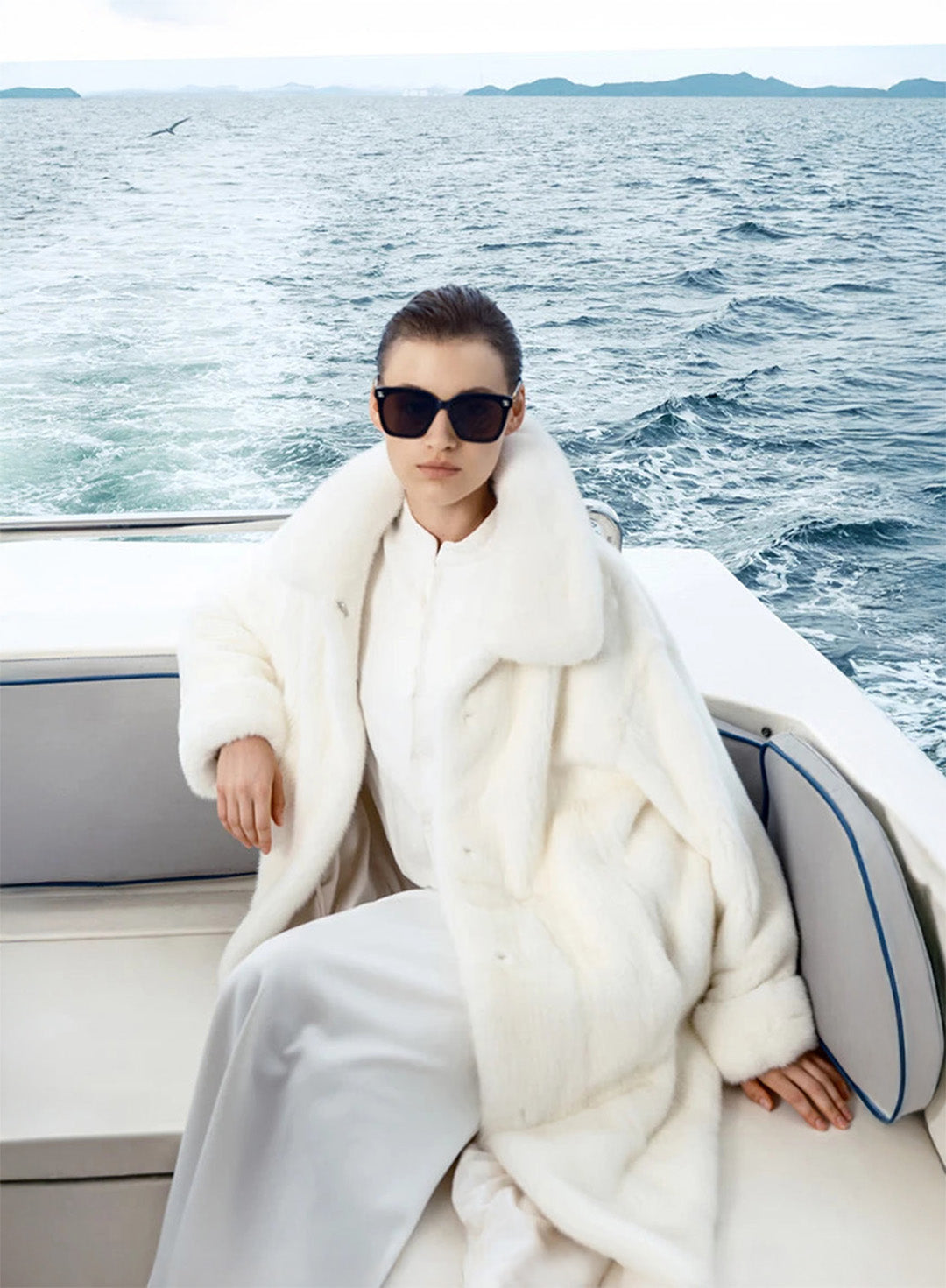 Women’s White Echo mink coat, model seated on yacht bench with one hand resting on the seat back, soft silhouette visible. — From Borcelleclothes.