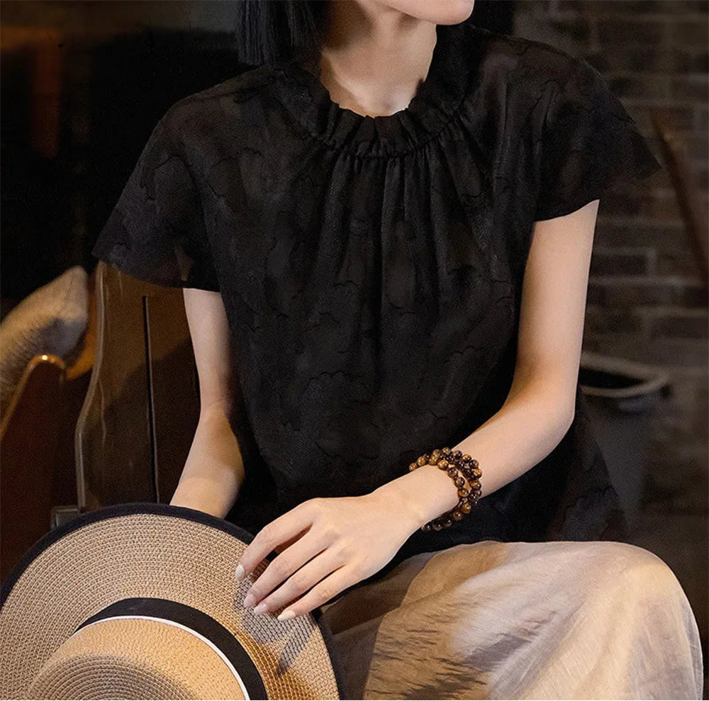 Women's Mulberry Silk Blouse in Bittersweet Noir — seated model holding a woven hat, highlighting the dimensional jacquard texture — From Borcelleclothes.