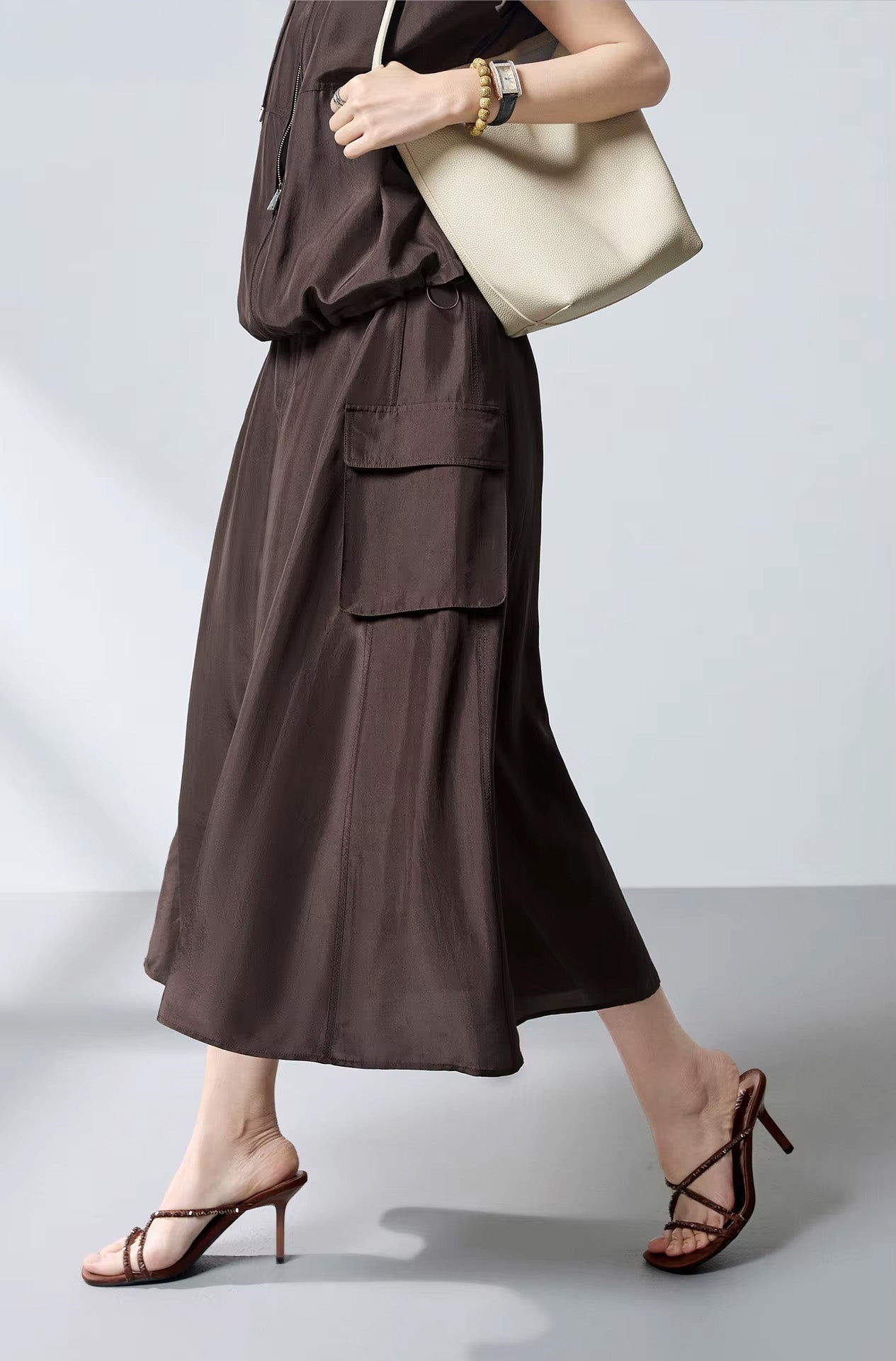 Side view of a model walking in a fluid Mulberry silk cargo skirt in Bitter Chocolate, carrying a tote bag over the shoulder — From Borcelleclothes.