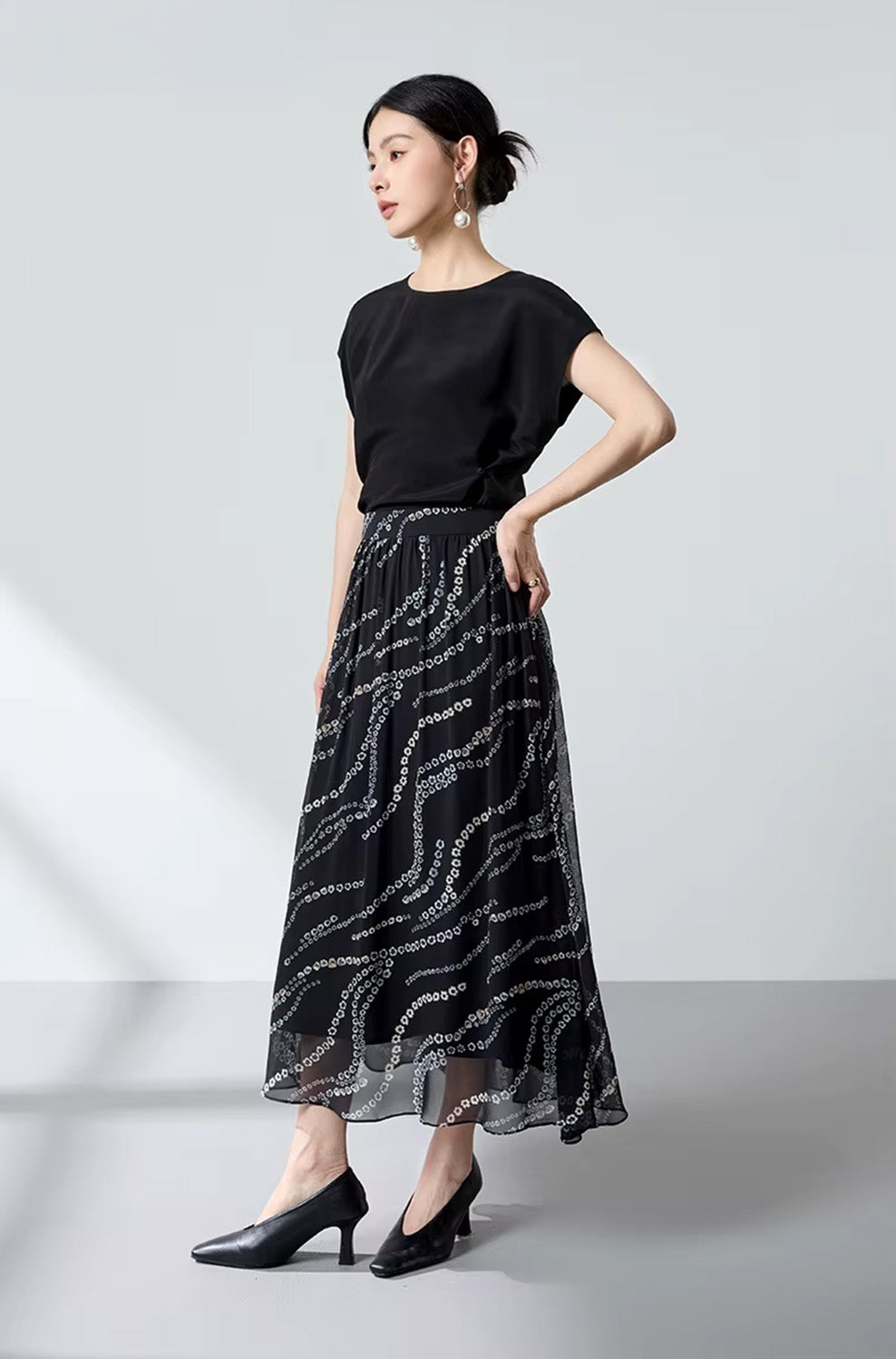 Full-height studio shot of a model with her hand on her hip, showcasing a flared black silk skirt with a white floral pattern — From Borcelleclothes.