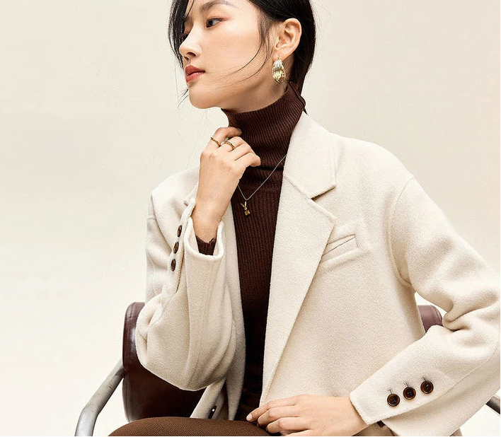 From Borcelleclothes: Close-up of a model in the Piano coat touching her collar of the turtleneck sweater, highlighting the clean, pure lines of the double-breasted silhouette.