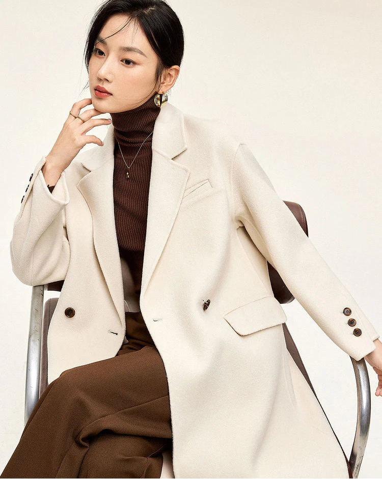 From Borcelleclothes: A seated model in the Piano ivory double-sided wool coat, thoughtfully touching her chin, highlighting the soft structure of the notched lapels.