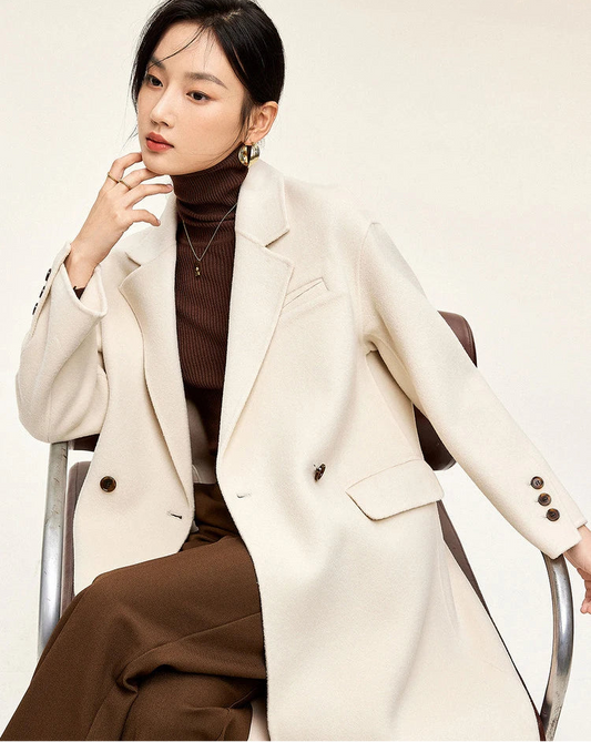 From Borcelleclothes: A seated model in the Piano ivory double-sided wool coat, thoughtfully touching her chin, highlighting the soft structure of the notched lapels.