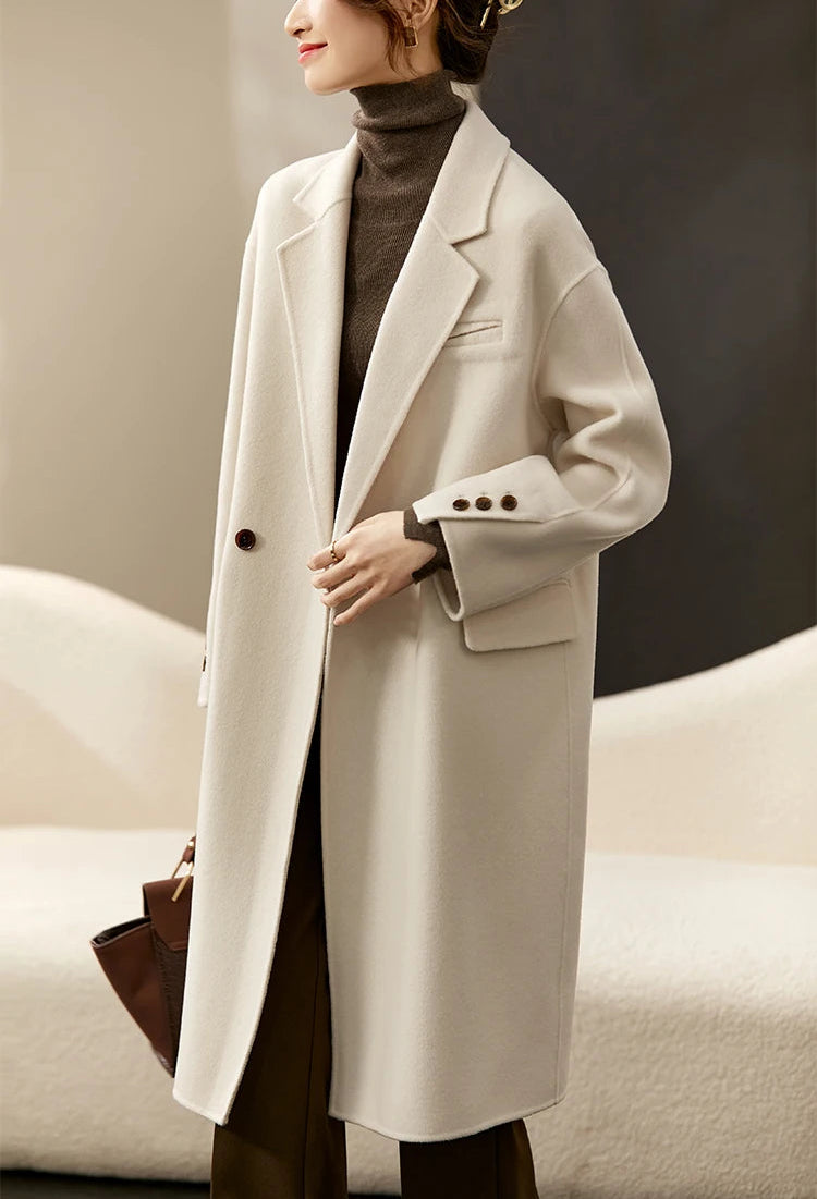 From Borcelleclothes: A model wearing the unfastened Piano coat, touching the front edge to reveal the artisanal invisible seams and the absence of a lining.