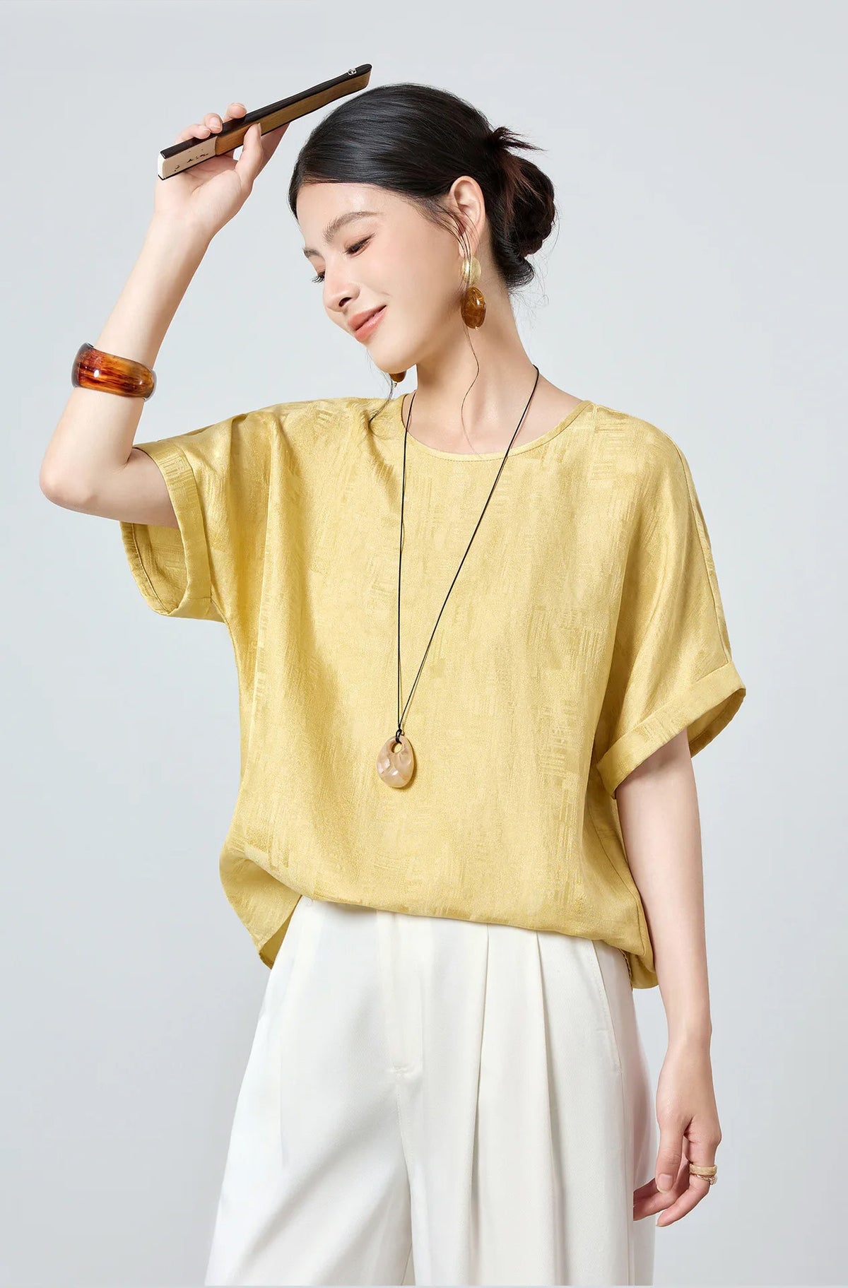 Editorial view of model in a Hakea Mulberry silk Sangbo jacquard top holding a decorative fan above her head — From Borcelleclothes.