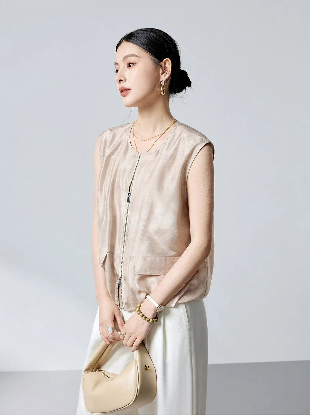 Front view of the model holding a bag with both hands in the sleeveless Mulberry Habotai silk blouson jacket in Whispering Wind — From Borcelleclothes.