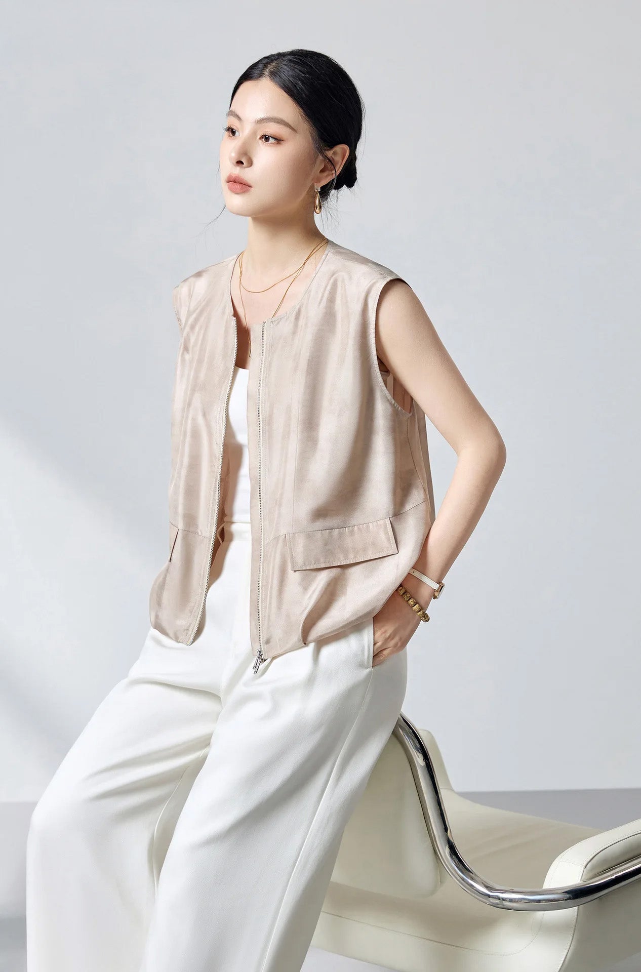 Model perching on the back of an armchair wearing the women's sleeveless Mulberry Habotai silk blouson jacket in Whispering Wind — From Borcelleclothes.