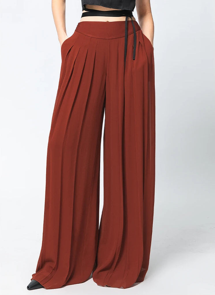 Two-layer pleated Mulberry silk georgette trousers in Contradictory Terracotta, model posing with hands in pockets — From Borcelleclothes.
