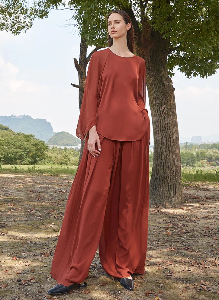 Model wearing two-layer pleated Mulberry silk georgette trousers in Contradictory Terracotta, standing outdoors in a natural setting — From Borcelleclothes.