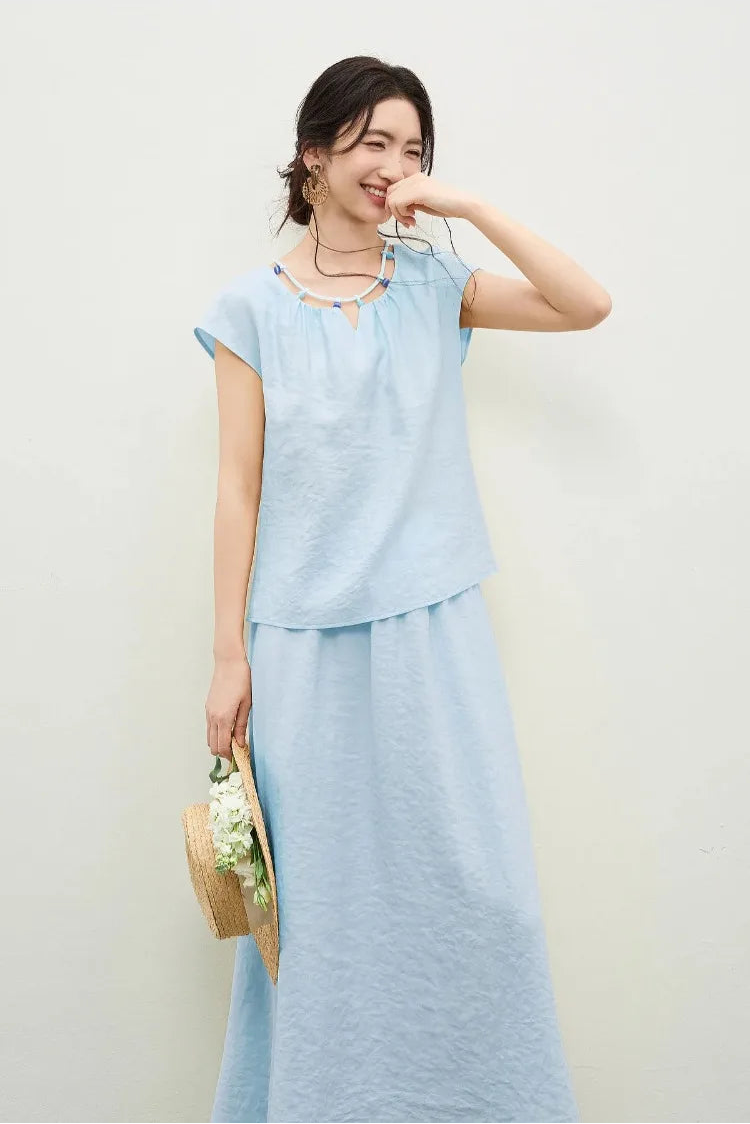 A woman in the sky blue viscose set with a gentle smile, touching her lips, framed by the creative beaded necklace detail — From Borcelleclothes.