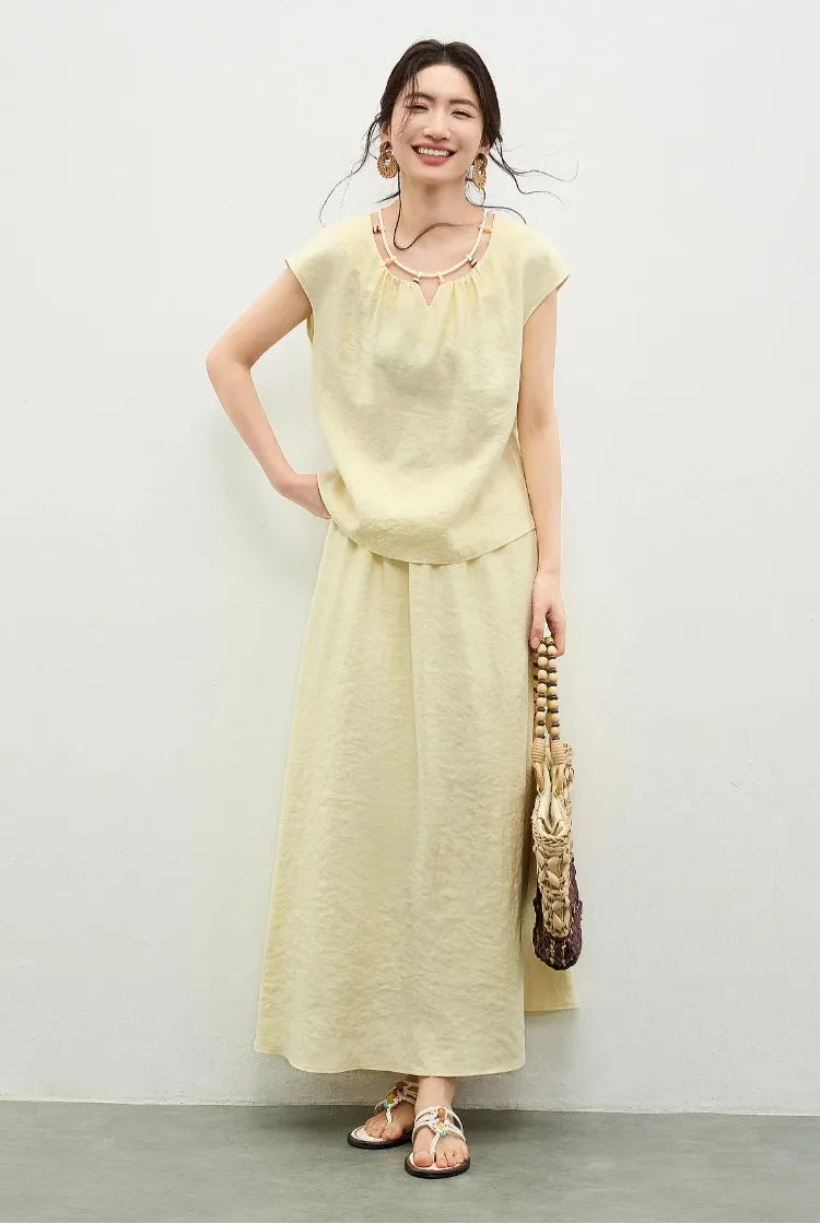 A smiling woman in a yellow viscose set, lightly touching the hem of her top to show the fluid drape of the fabric — From Borcelleclothes.