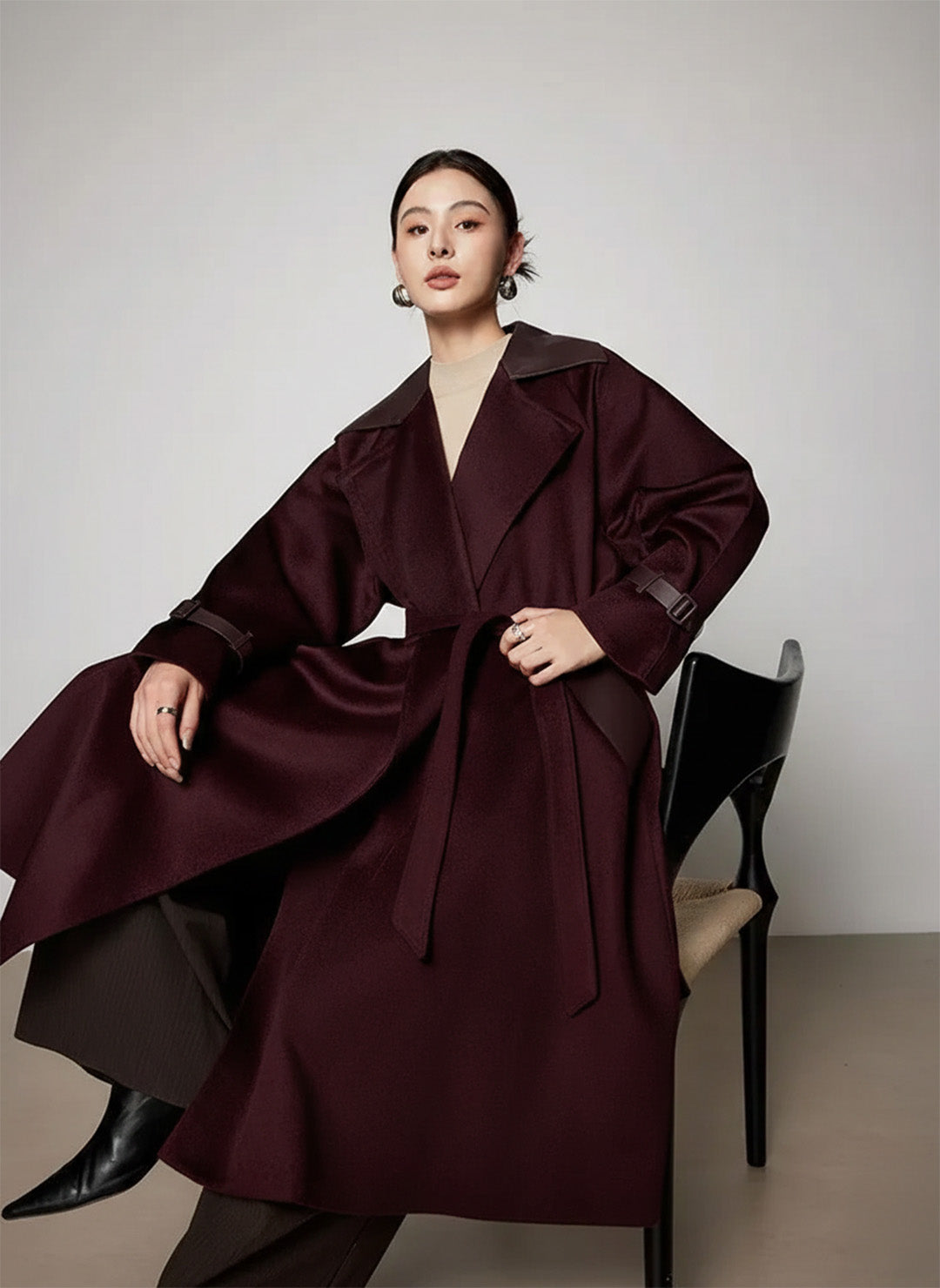 Model sitting in a chair wearing the Bordeaux wool coat, showcasing how the fabric drapes and the sheepskin collar's texture. From Borcelleclothes.