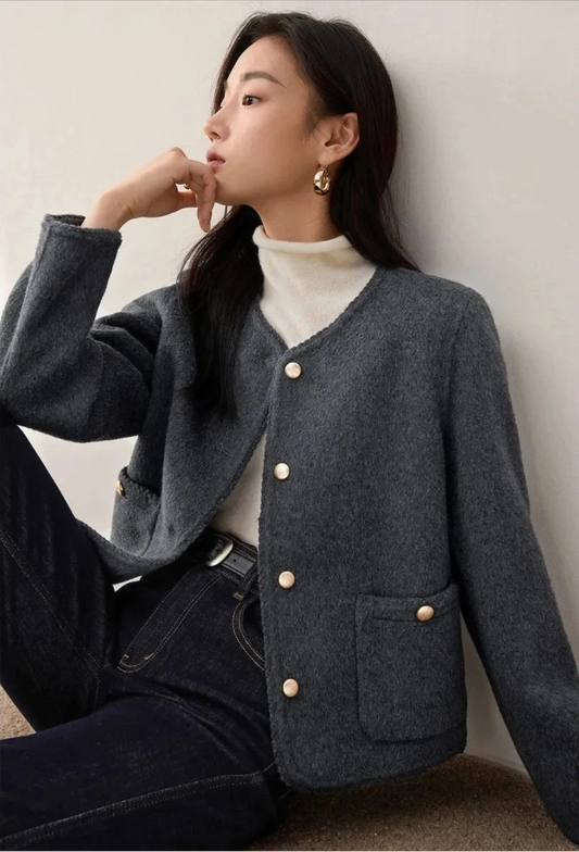 A woman in a Perle Noir double-sided wool jacket, seated in a thoughtful pose with her hand to her chin, showcasing the elegant fit and soft texture. — From Borcelleclothes.
