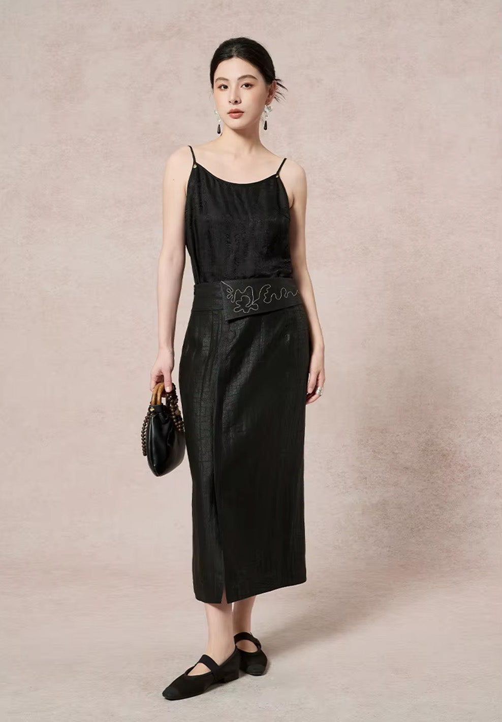 Full-length front view of a model in a black asymmetric Xiangyunsha silk midi skirt, showcasing the decorative placket and front vent while holding a bag — From Borcelleclothes.