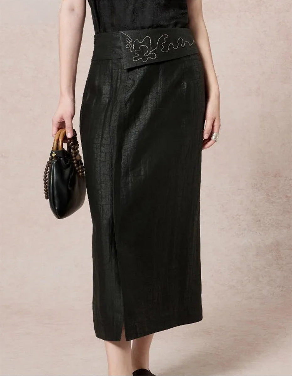 Close-up of a model wearing a structured black Xiangyunsha silk midi skirt with an embroidered overlay waist panel, holding a handbag — From Borcelleclothes.