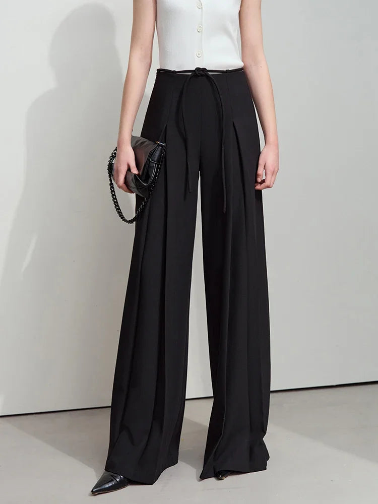 Model holds a bag while wearing Onyx-colored trousers with deep pleats and minimalist structure. From Borcelleclothes.