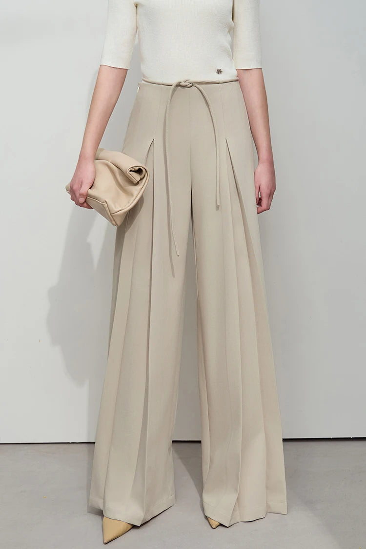Model holds a structured bag while wearing draped trousers in Silk colors with sculptural pleats. From Borcelleclothes.