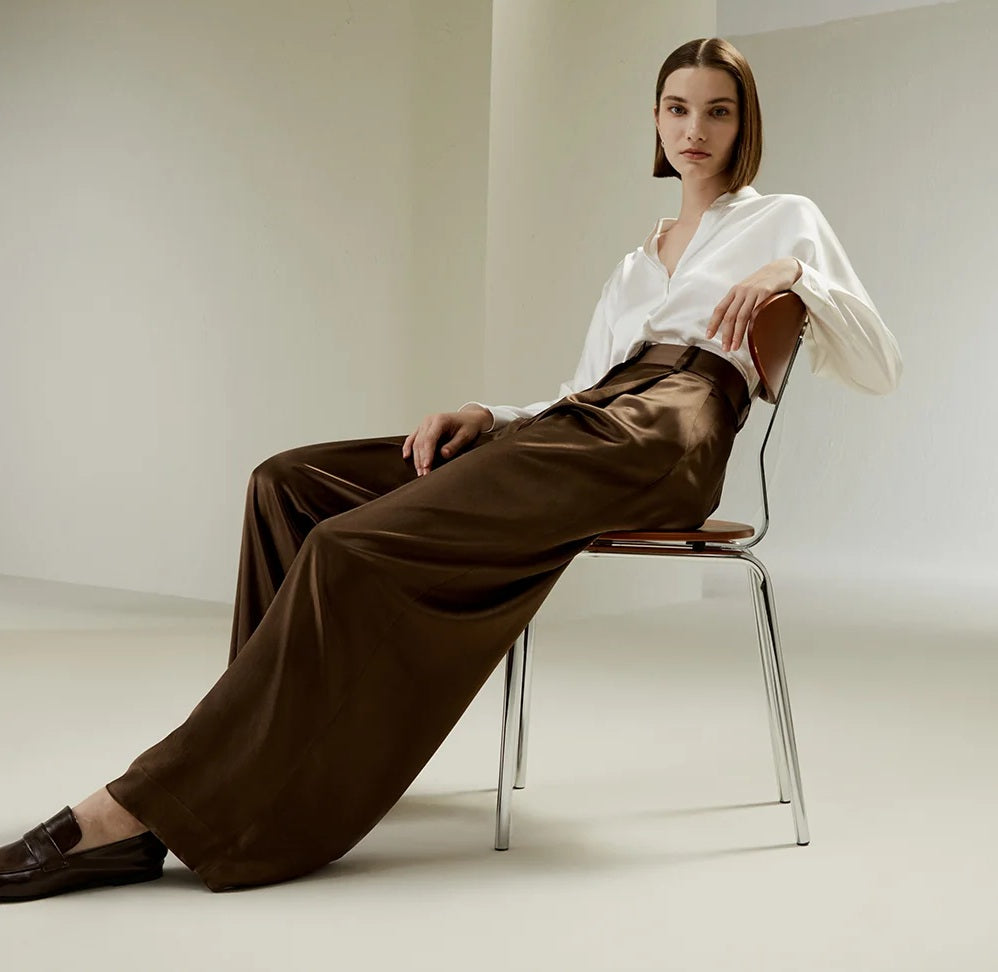 Model in Cafe Noir silk trousers reclining on a chair, illustrating the fluidity and richness of the fabric. From Borcelleclothes.