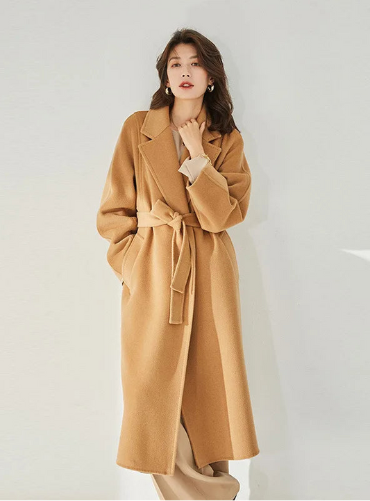 A model wearing the Women's Wrap Coat Redefined in Brown Sugar, gently touching the collar, emphasizing its refined notch lapels and tailored elegance. From Borcelleclothes.